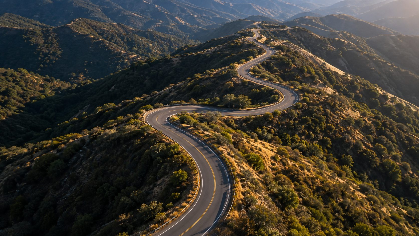 Aerial view of a winding two-lane mountain road at golden hour.